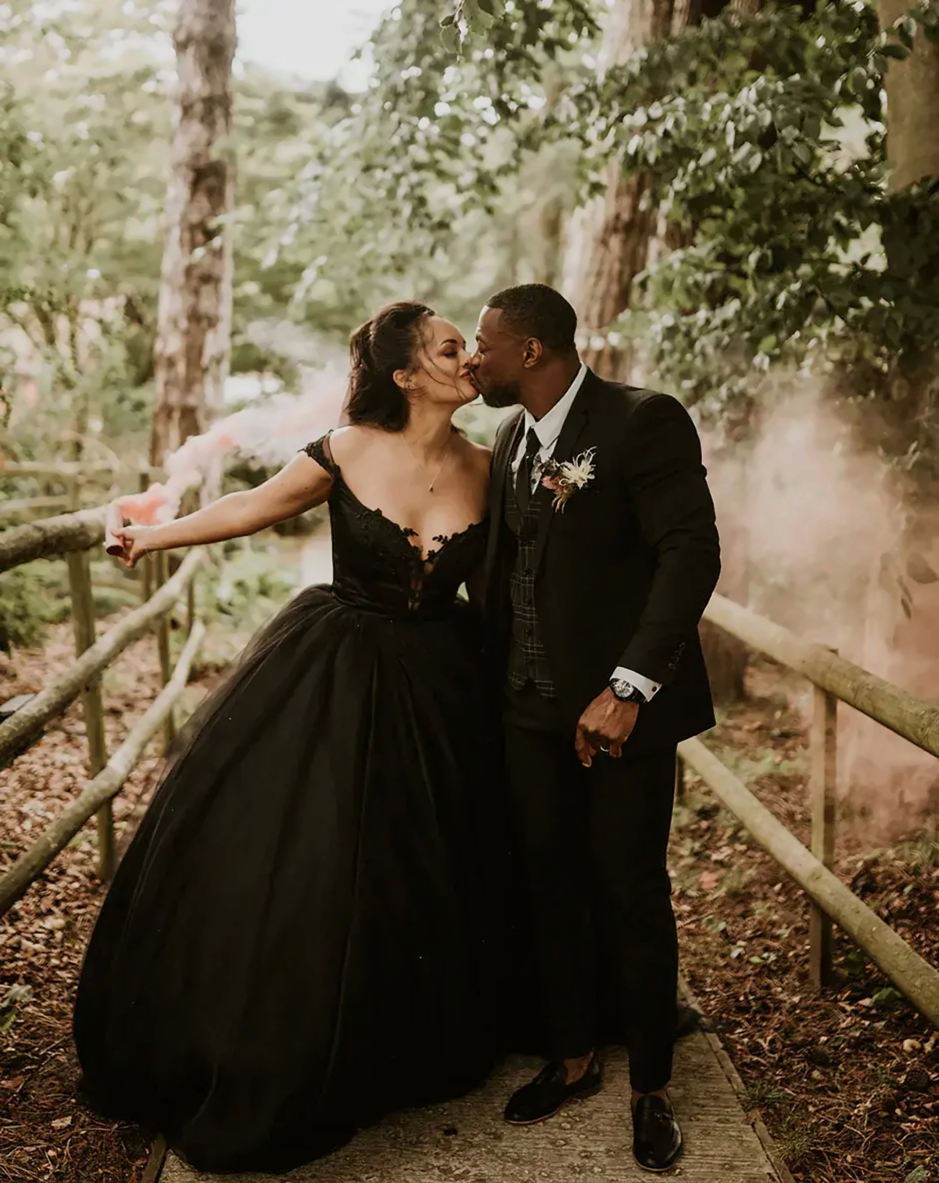 A romantic outdoor wedding scene featuring a bride and groom sharing a kiss. The bride is wearing a dramatic off-the-shoulder black gown with a full, voluminous skirt and lace bodice, while the groom is dressed in a sharp black suit with a boutonniere. They stand on a wooded path surrounded by greenery, with the bride holding a pink smoke stick that adds a whimsical, ethereal atmosphere to the scene. The couple is framed by the natural beauty of the trees, with soft lighting adding to the enchanting, intimate mood of the moment.