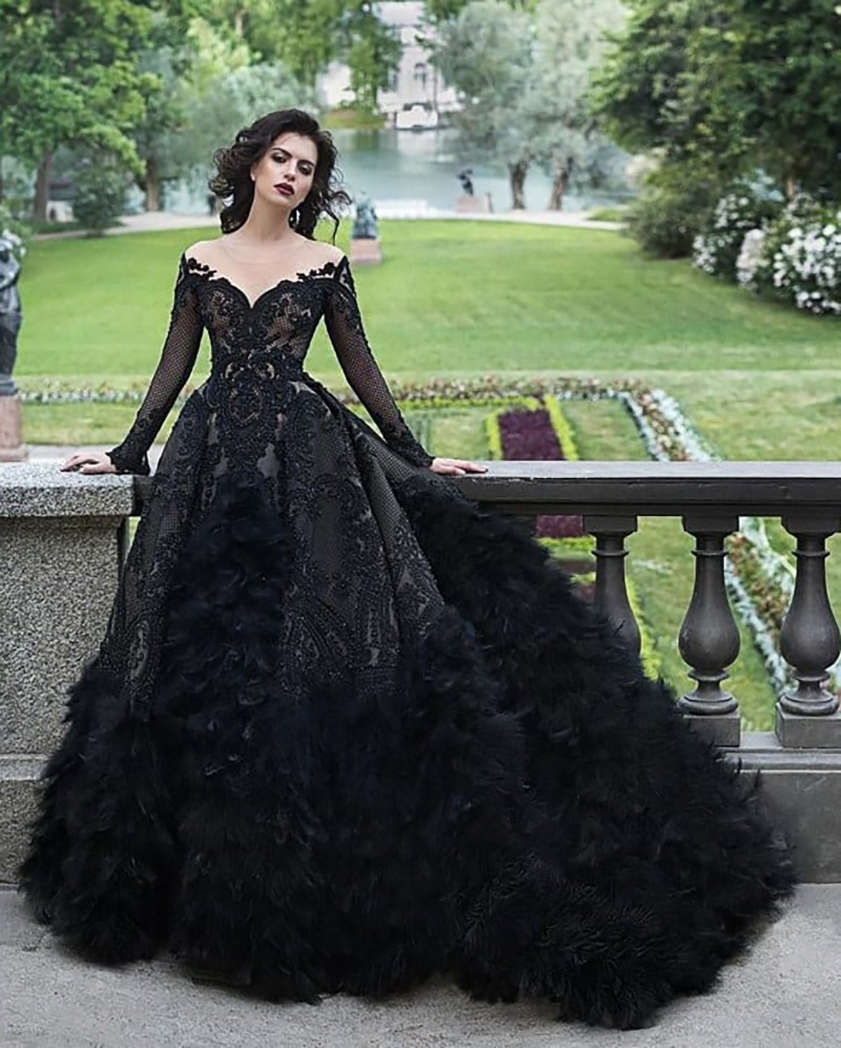 Una mujer de aspecto majestuoso vestida con un dramático vestido negro con los hombros descubiertos de pie en un jardín formal. El vestido presenta intrincados detalles de encaje en el corsé y una falda voluminosa con capas texturizadas parecidas a plumas en la parte inferior. Su cabello oscuro está peinado en rizos sueltos y su expresión es tranquila y confiada. El fondo es un jardín bellamente cuidado con exuberante vegetación, flores y un río tranquilo a lo lejos, creando una atmósfera refinada y elegante.