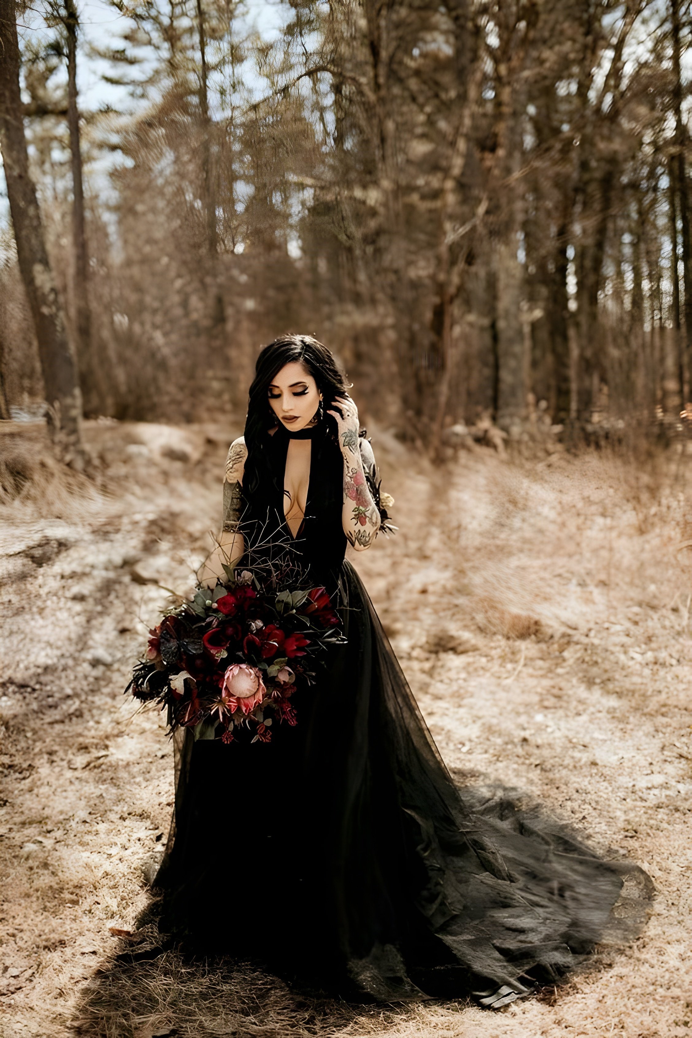 Una mujer está sola en un bosque, vestida con un dramático vestido negro con un profundo escote y una falda fluida. Sostiene un gran ramo lleno de flores rojas oscuras y negras. Su largo cabello oscuro cae en cascada sobre sus hombros y sus brazos presentan tatuajes visibles, lo que añade un toque atrevido a su apariencia. El fondo del bosque, con árboles altos y colores tenues, crea una atmósfera serena pero ligeramente gótica, complementando la audacia de su atuendo y ramo.