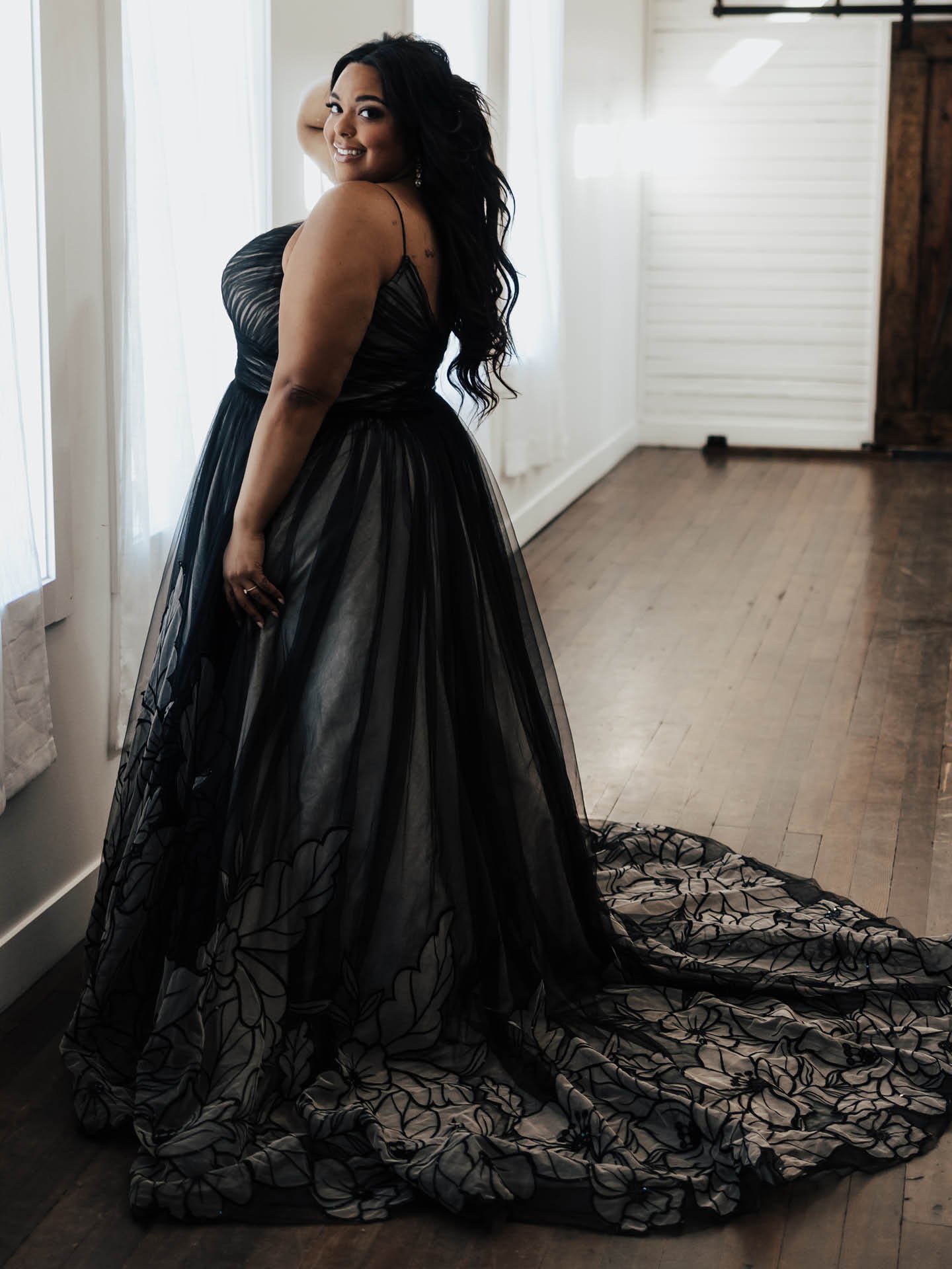 Una mujer está de pie en una habitación suavemente iluminada, vestida con un impresionante vestido negro con una falda de tul fluida y detalles florales a lo largo del dobladillo y la cola. El vestido presenta finos tirantes y un corsé fruncido que favorece su figura. Sonríe con confianza por encima de su hombro, con su largo cabello oscuro cayendo en cascada por su espalda. La habitación tiene suelos de madera, paredes blancas y grandes ventanas, creando una atmósfera luminosa y aireada que contrasta maravillosamente con su elegante vestido oscuro.