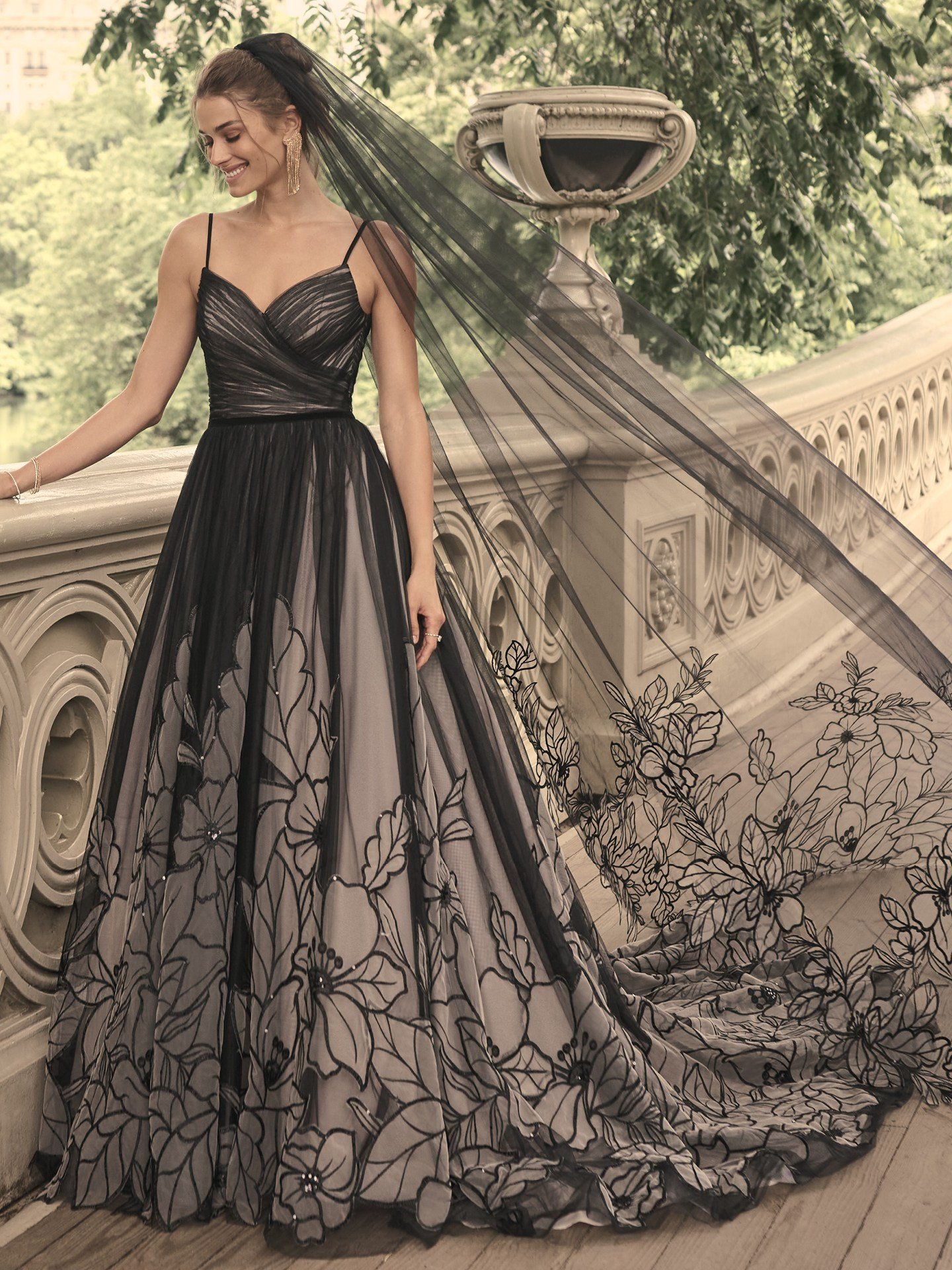 A smiling woman stands outdoors on a stone terrace, wearing a flowing black and gray gown with thin straps and a ruched bodice. The skirt features large, intricate floral patterns that extend onto the gown’s long train. She wears a sheer black veil that drapes down her back and catches the light breeze. The background shows greenery and an ornate stone railing, adding a romantic, timeless feel to the image.