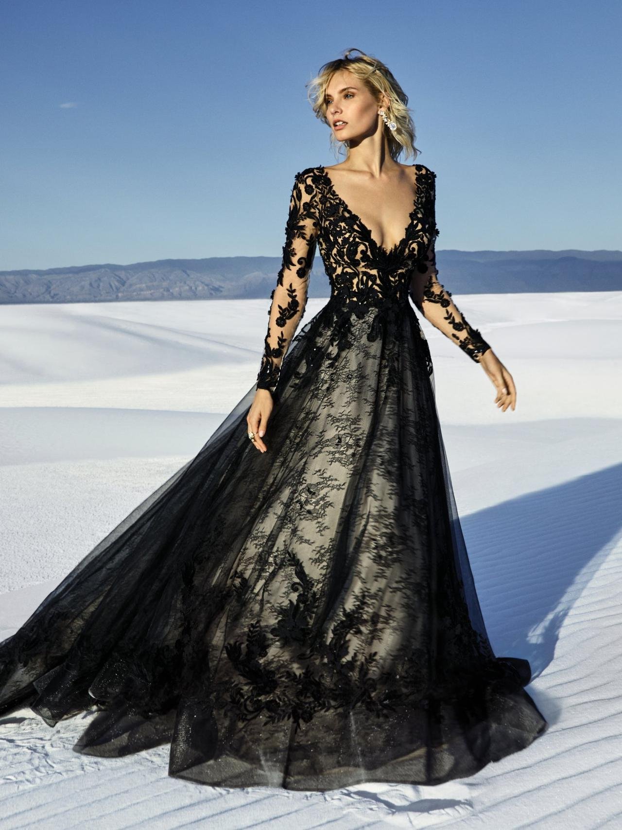 A woman in a black lace and tulle gown with a deep V-neckline and long sheer sleeves stands in a desert landscape with white sand dunes. The intricate black floral appliqué on her gown contrasts with the bright sky and sand. Her blonde hair is loosely styled, and she wears statement earrings, exuding elegance against the stark, natural background.