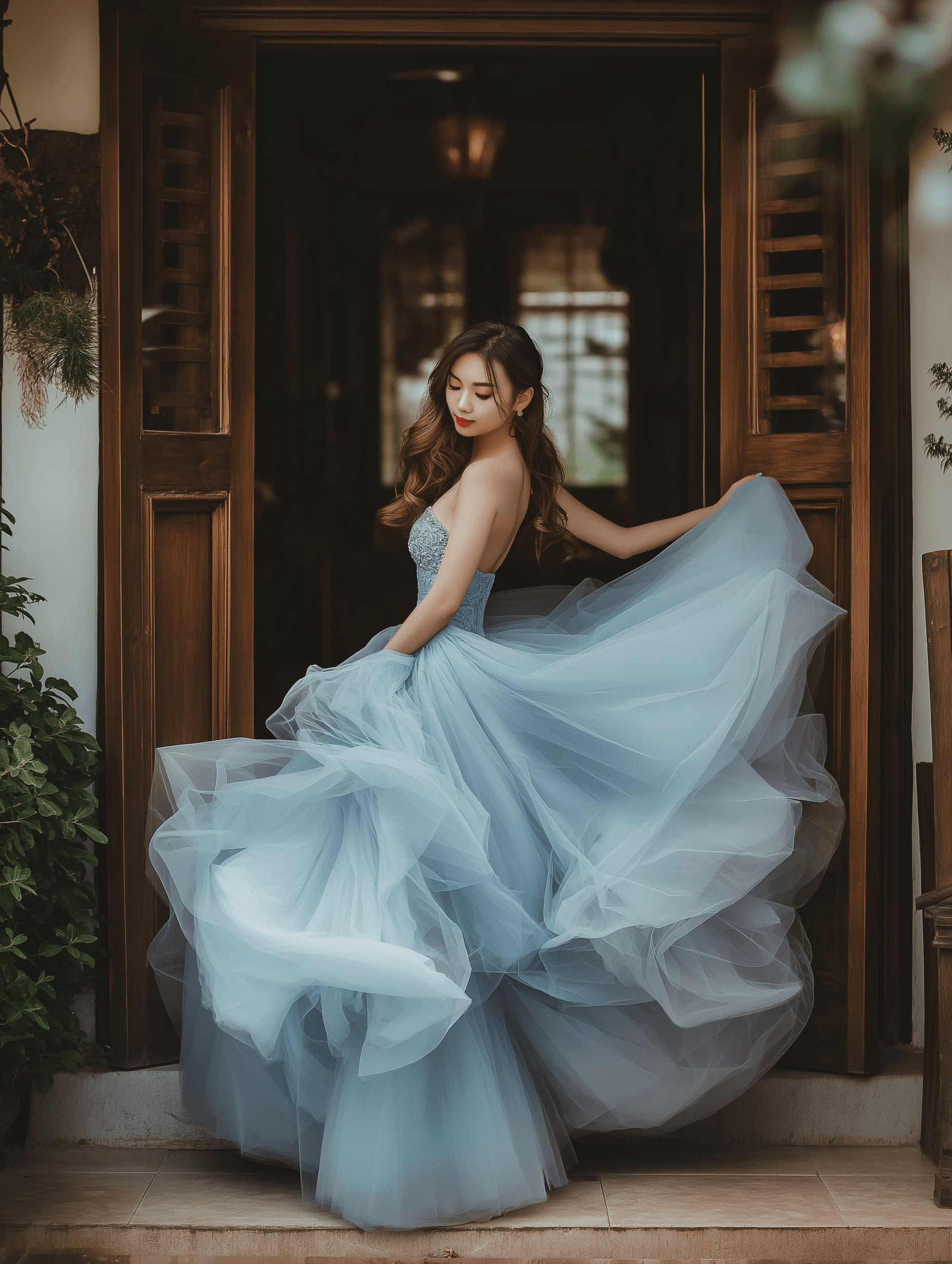 A woman wearing a light blue strapless gown with a voluminous tulle skirt stands in a doorway, gracefully twirling the fabric of her dress. Her hair is styled in soft waves, and she looks down gently as the layers of her gown billow around her. The setting is a cozy indoor-outdoor environment framed by wooden doors.