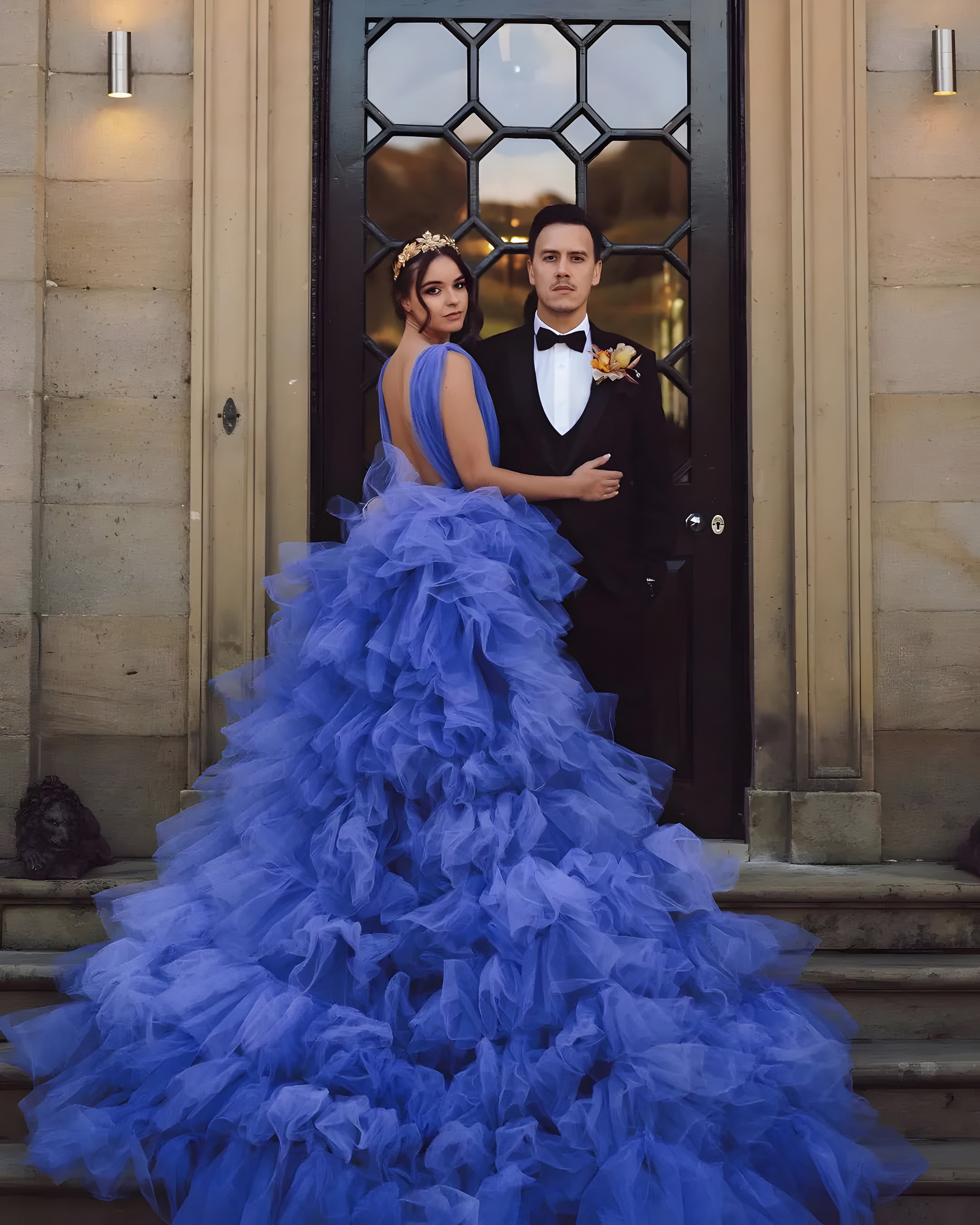 A woman in a vibrant royal blue gown with voluminous ruffled layers stands beside a man in a black tuxedo. They are positioned in front of a grand black door with glass panels. The woman’s dress has a dramatic, flowing train and she wears a gold floral headpiece, while the man wears a bow tie and a boutonnière.