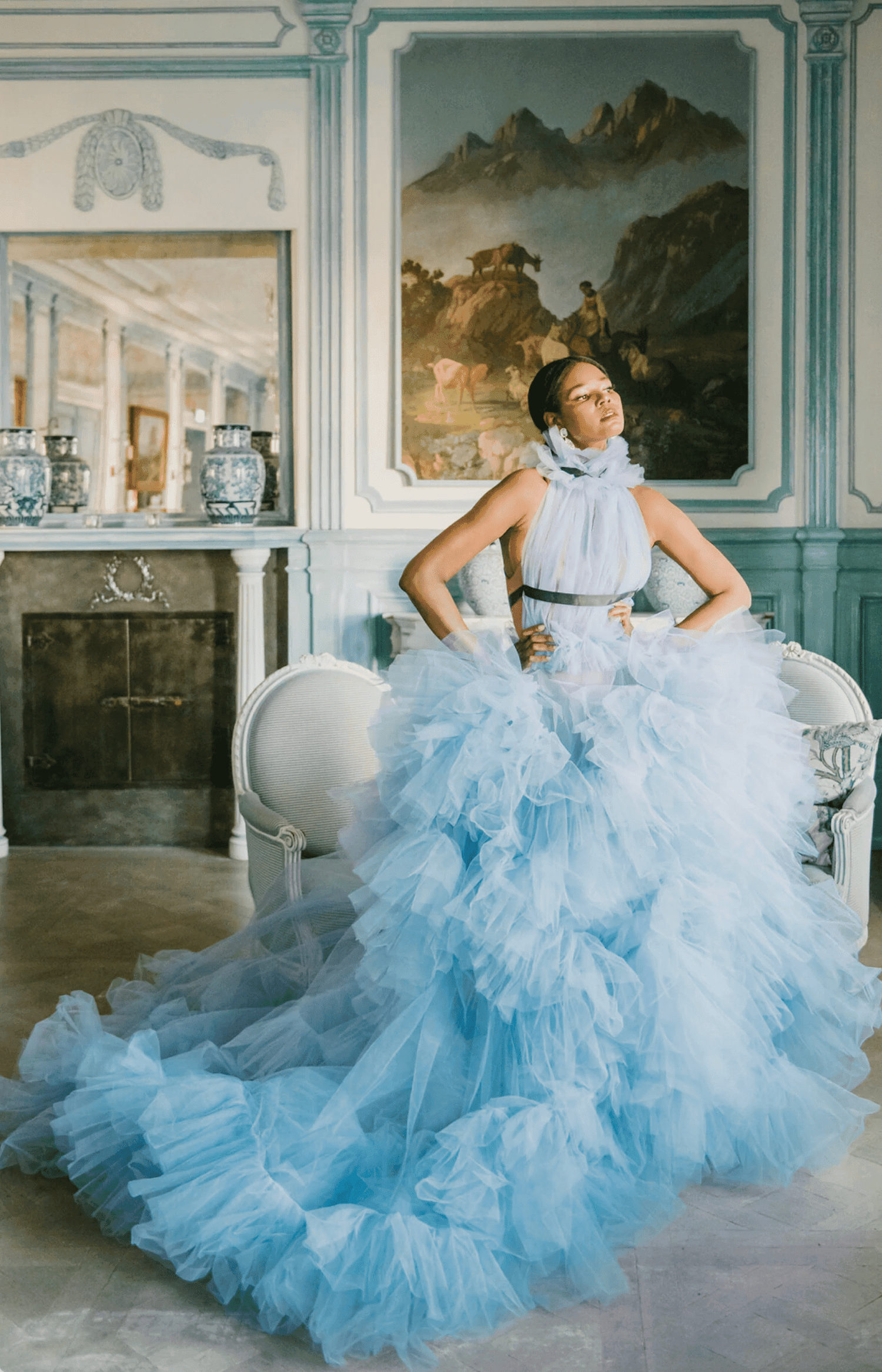 A woman in a voluminous light blue tulle gown stands confidently in a luxurious, vintage-style room with classical paintings and antique furniture. The gown features layers of ruffled fabric and a high neckline, cinched at the waist with a dark belt. The opulent surroundings enhance the ethereal, elegant feel of the scene.