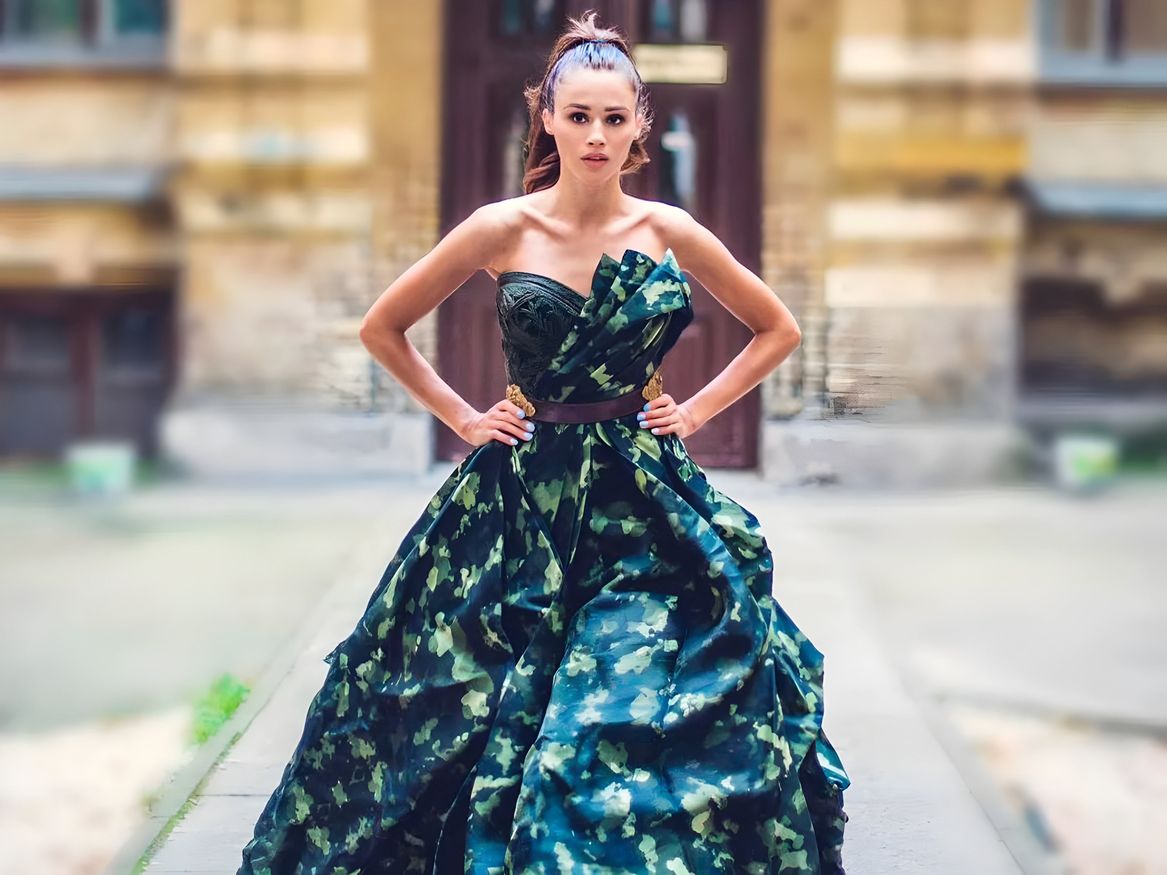 A woman stands confidently wearing a dark green and black strapless gown with bold, abstract patterns. The dress features a dramatic, voluminous skirt and an asymmetrical bodice with artistic draping. Her hair is styled in a high ponytail as she poses outdoors in an urban setting.