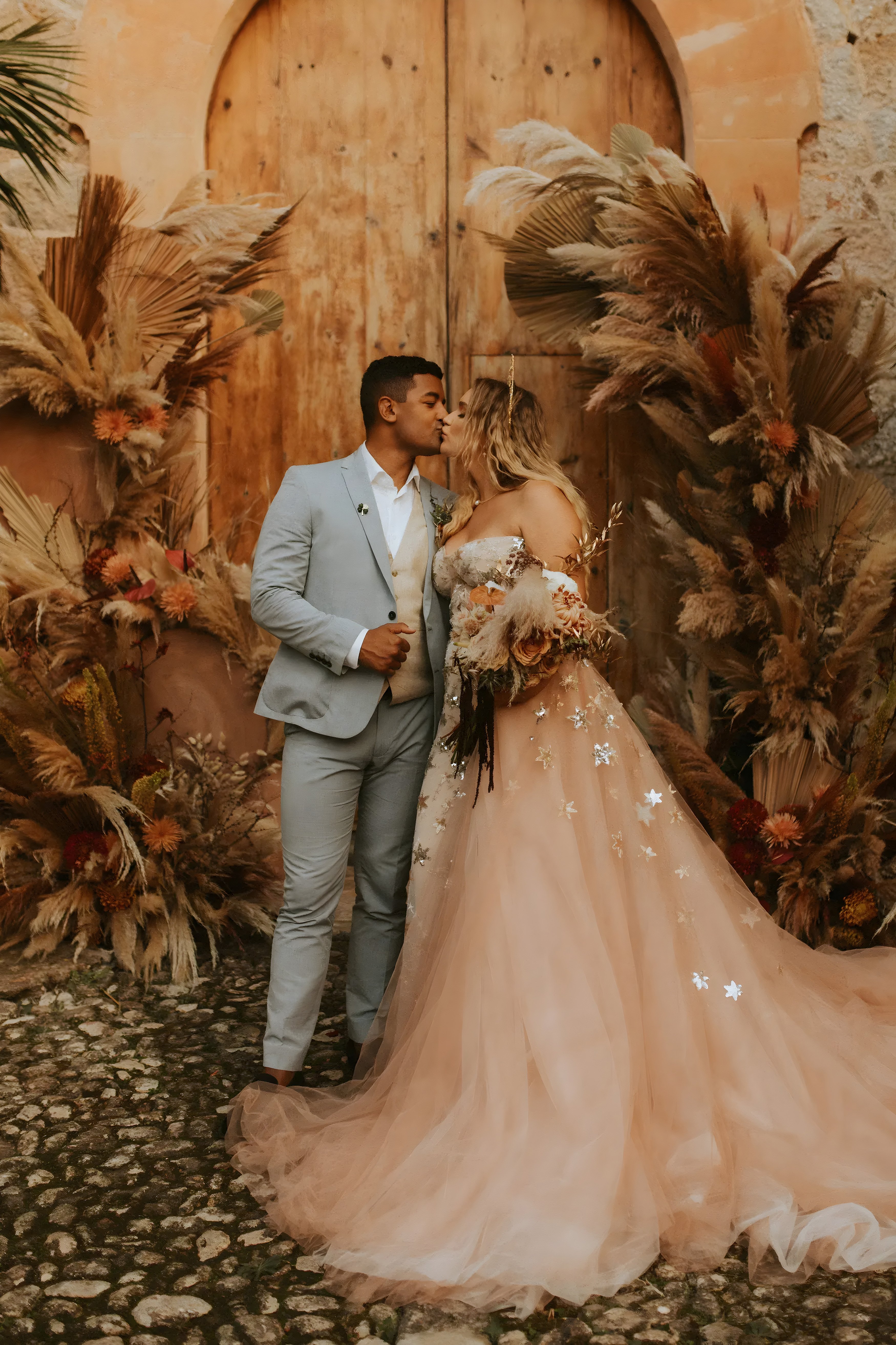 A couple is sharing a kiss in front of a rustic wooden door, surrounded by earthy, dried floral arrangements. The bride is wearing an elegant off-the-shoulder blush gown adorned with sparkling floral appliqués, and the groom is dressed in a light gray suit with a cream-colored vest underneath. The setting has warm, neutral tones, with pampas grass and dried flowers framing the couple.