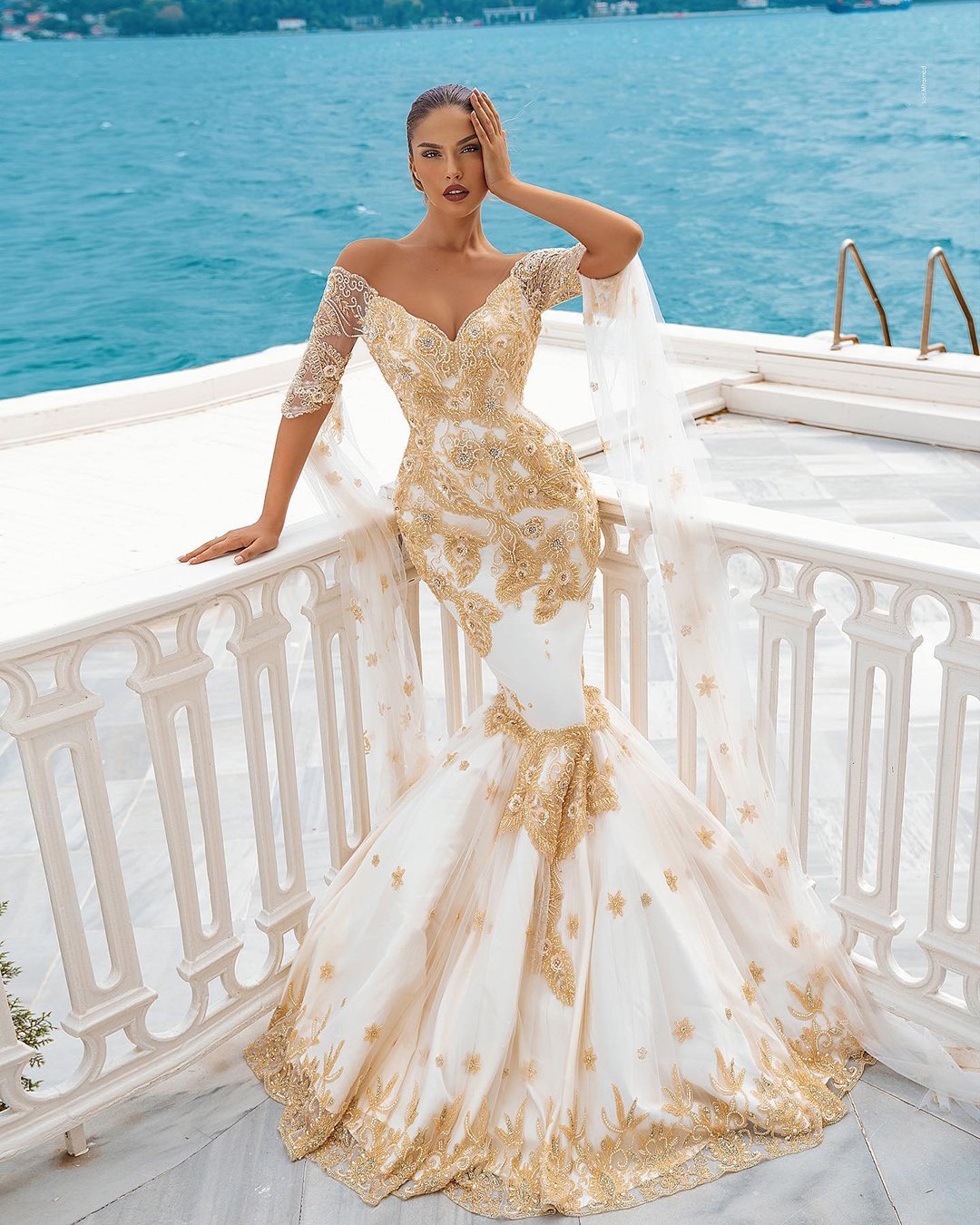 A woman is standing on a white balcony by the sea, wearing a fitted mermaid-style gown with gold lace embroidery and off-the-shoulder sleeves. The gown features a flowing sheer cape adorned with gold details. She has one hand on her face and the other resting on the railing, with the ocean and distant shoreline in the background.