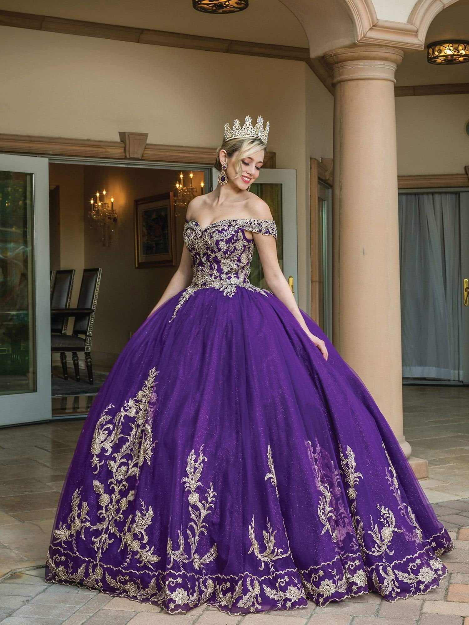 A woman is standing outdoors in a royal purple, off-the-shoulder ball gown adorned with intricate gold embroidery. She is wearing a large, ornate crown and smiling as she looks down. The setting is a grand entrance to a building, with stone pillars and elegant lighting visible inside.