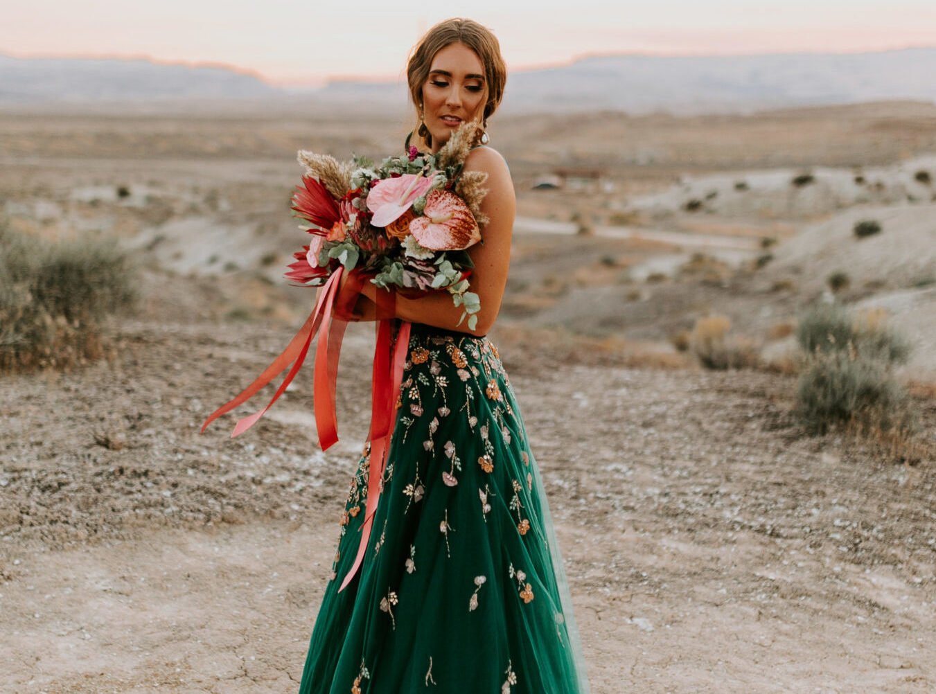 Una mujer está de pie en un paisaje desértico, vistiendo un vestido verde con bordados florales. Sostiene un gran y colorido ramo de flores con cintas rojas y rosas que flotan en el viento. El fondo muestra tierras áridas y colinas distantes, con luz suave del sol iluminando la escena.