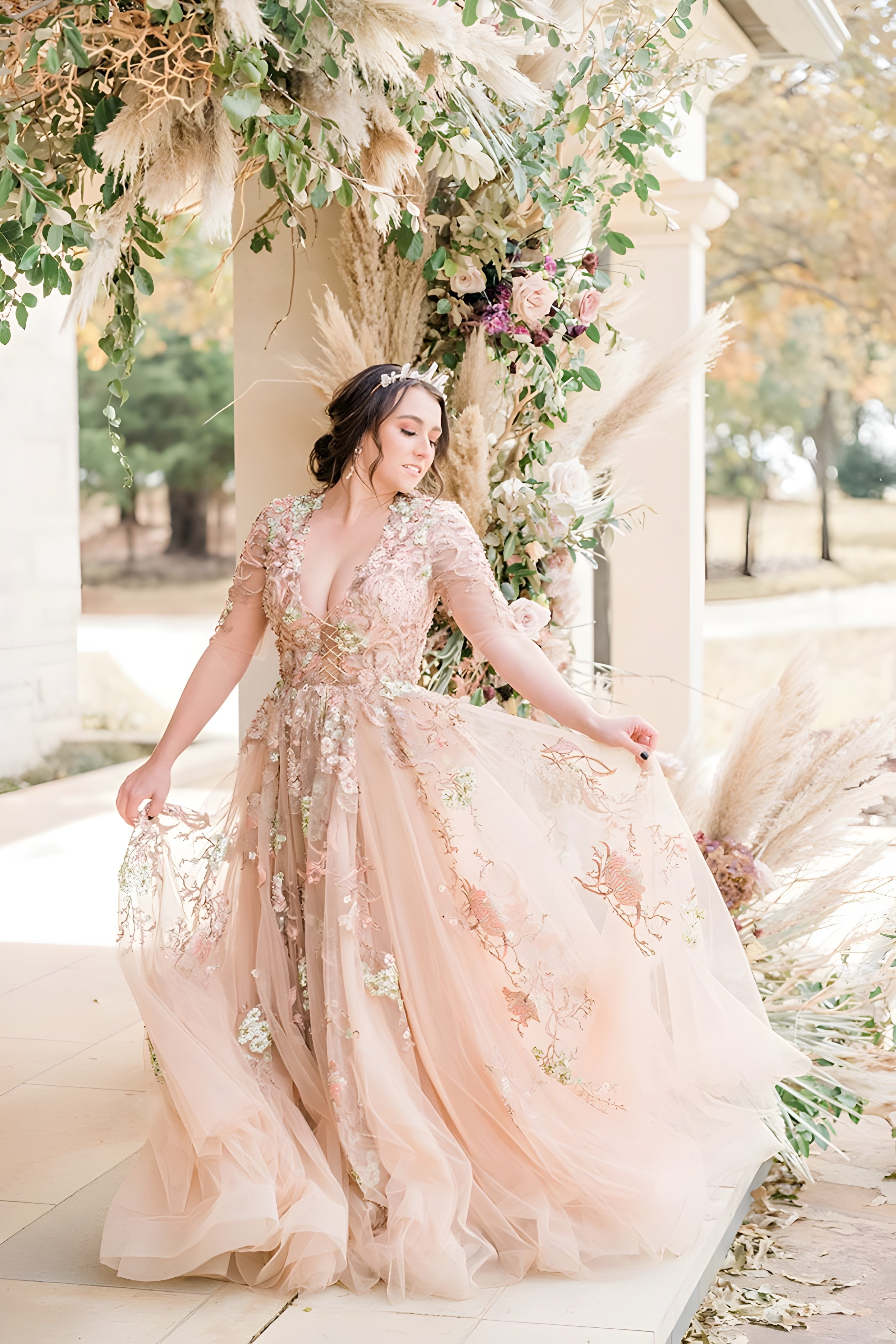 A woman is standing in a flowing, blush-colored gown with intricate floral embroidery. She is posing under a decorated archway with pampas grass, greenery, and roses, gently holding the edge of her dress. The background includes a bright outdoor setting with soft natural light.