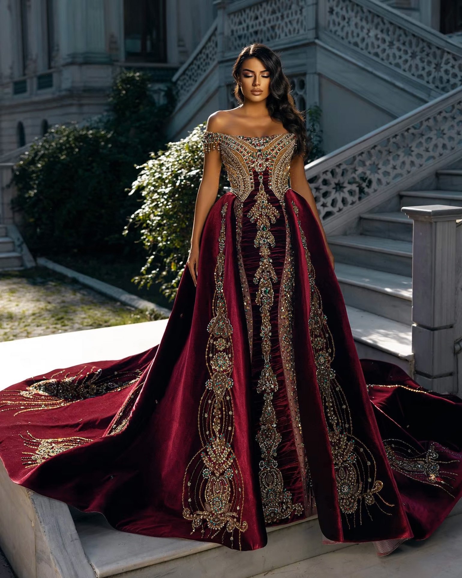 A woman stands on a staircase wearing an opulent off-the-shoulder burgundy gown adorned with intricate gold and jewel-encrusted embroidery. The dress has a dramatic train and a regal, structured design. The background includes a marble staircase and a lush garden, adding to the luxurious atmosphere.