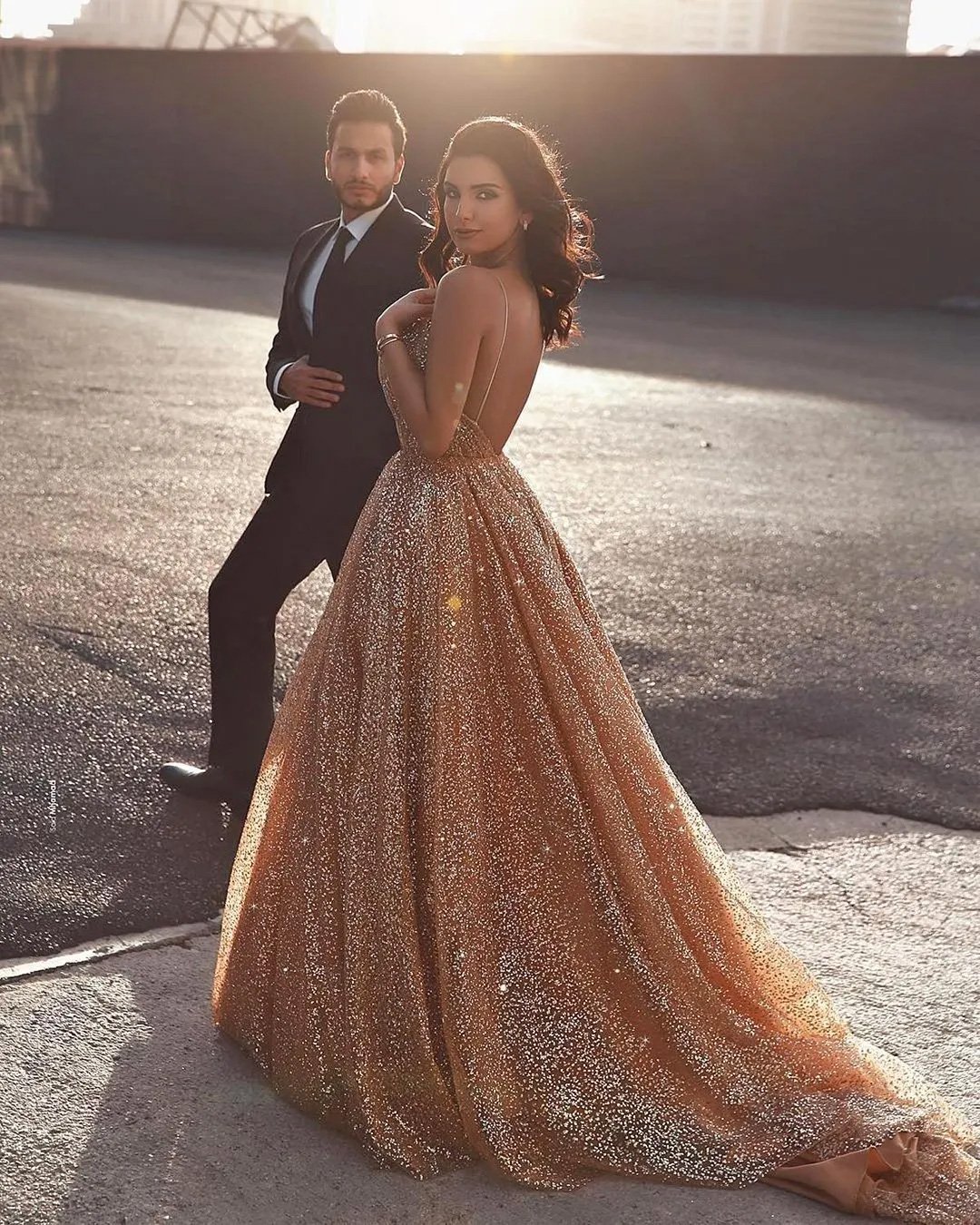 Une femme vêtue d'une robe à paillettes dorée, longue comme le sol et dos nu, se tient gracieusement dans un décor urbain à l'heure dorée. Ses cheveux noirs tombent en vagues et elle regarde l'appareil photo par-dessus son épaule. Un homme en smoking noir se tient derrière elle, tous deux baignés par la lumière chaude du soleil, ajoutant une touche glamour et élégante à la scène.