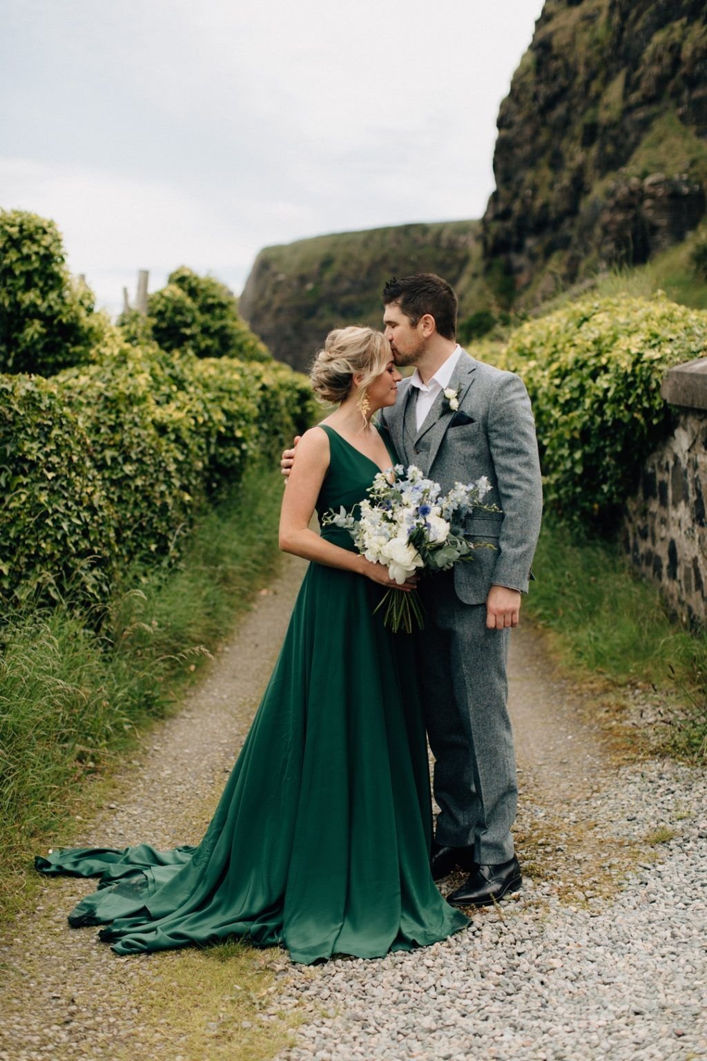 Un couple se tient sur un chemin de gravier entouré de verdure, l'homme embrassant le front de la femme. Elle porte une robe vert émeraude profond avec une traîne fluide et tient un bouquet de fleurs blanches et bleues, tandis que lui porte un costume en tweed gris. La scène se déroule dans un paysage naturel luxuriant, avec des falaises en arrière-plan, évoquant un moment paisible et romantique.