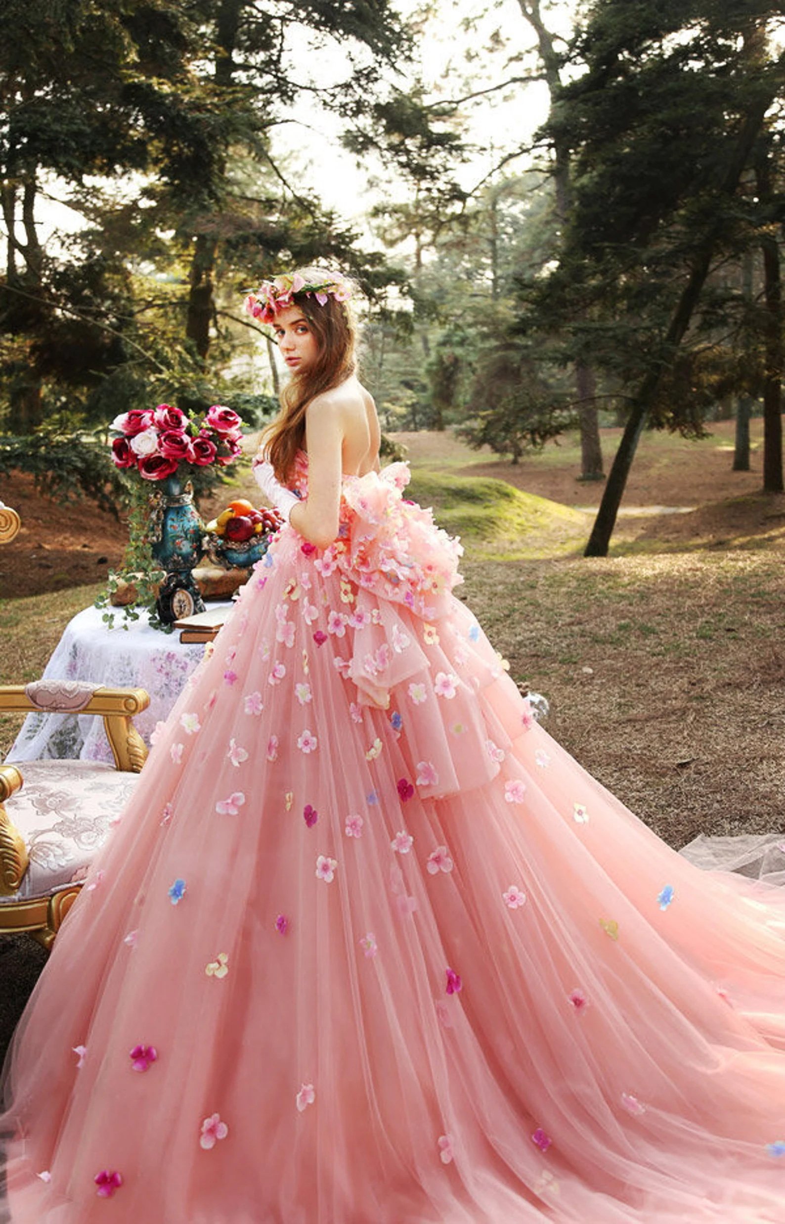 A woman stands outdoors in a pastel pink wedding dress adorned with colorful floral appliqués. She wears a flower crown and looks over her shoulder. The dress has a full, flowing skirt and is set in a forested area with a table decorated with flowers and fruit.