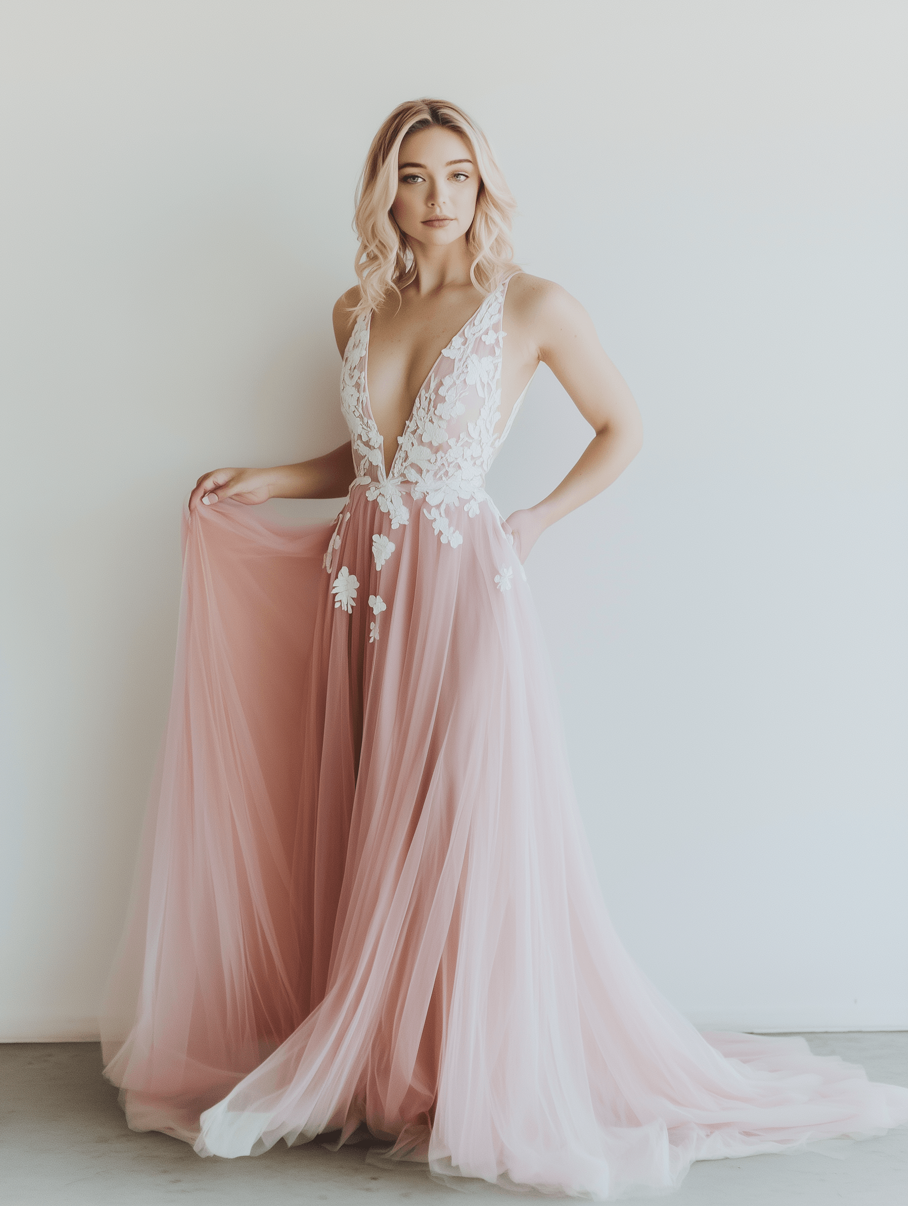 Una donna si trova in un abito da sposa rosa chiaro con un profondo scollo a V e applicazioni in pizzo floreale sul corpetto. L'abito ha una gonna in tulle fluente e lei tiene delicatamente parte dell'abito con una mano mentre l'altra mano è appoggiata sulla vita. I suoi capelli sono acconciati in onde morbide e lei si trova davanti a uno sfondo bianco semplice.