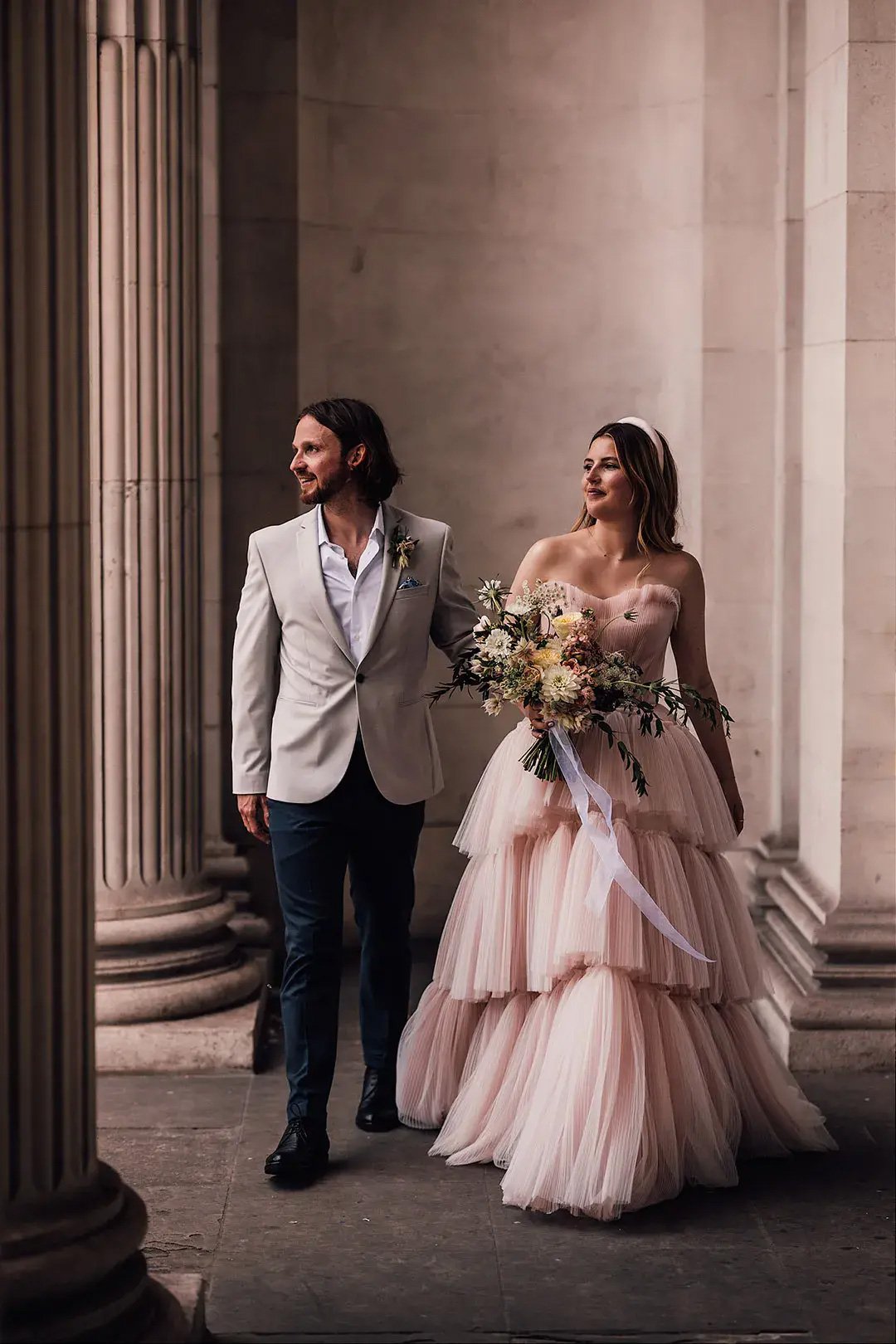 Uma noiva em um vestido de noiva de tule rosa blush em camadas segura um buquê e está ao lado do noivo, que veste um blazer de cor clara e calças escuras. Eles estão caminhando juntos em um cenário elegante com colunas e iluminação suave.