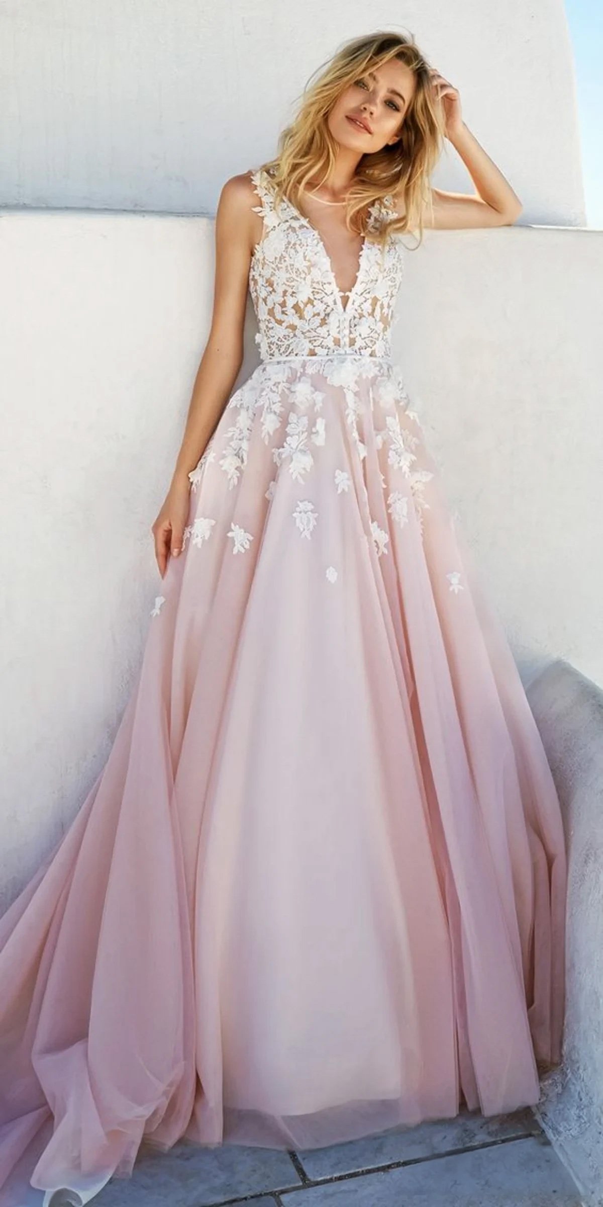 Une femme portant une robe de mariée rose blush avec un corsage en dentelle et des appliqués floraux se tient devant un mur de couleur claire. La robe a un décolleté en V plongeant, des détails en dentelle transparente, et une jupe en tulle volumineuse.