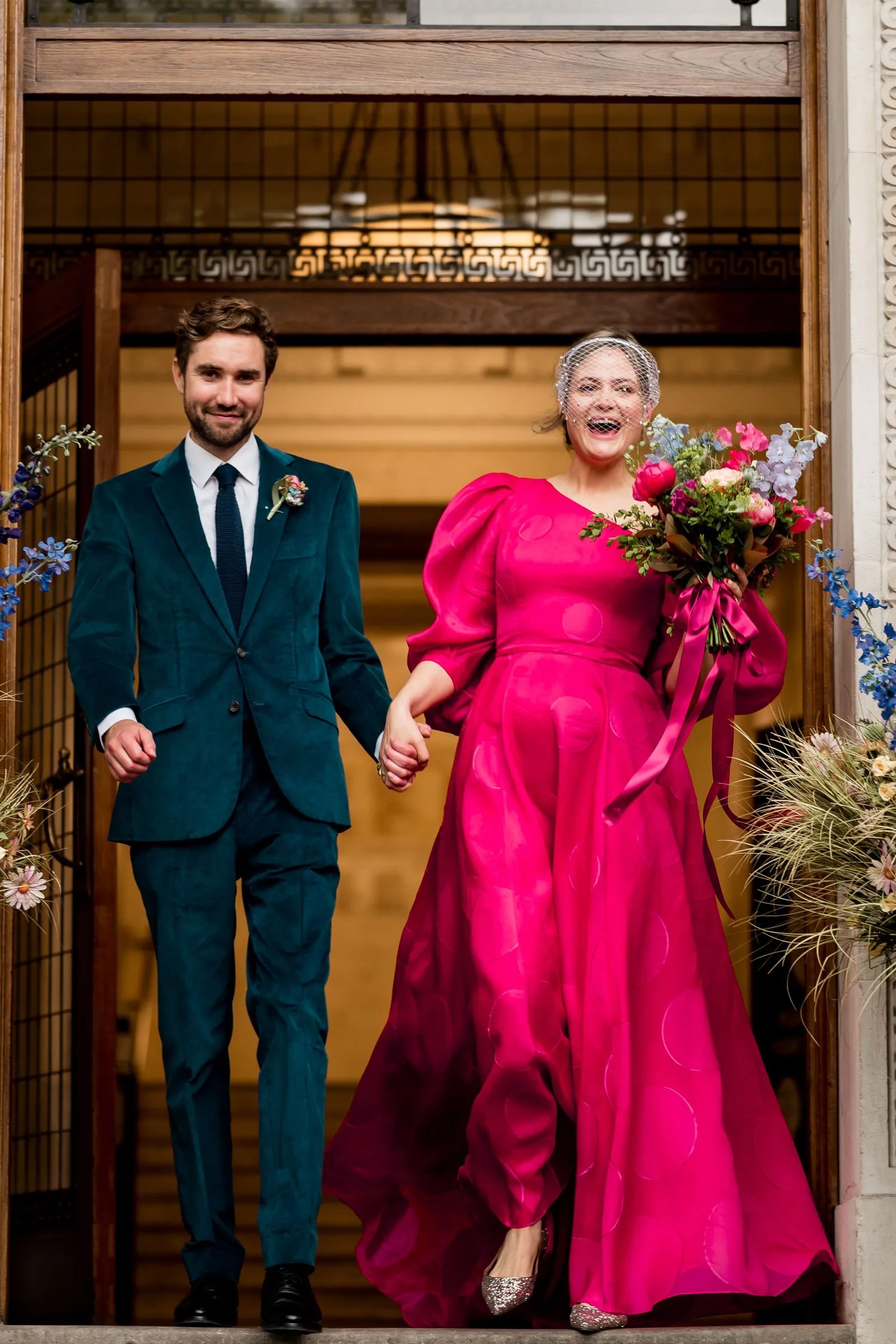 A bride in a vibrant pink wedding dress with puffed sleeves and a playful silhouette holds a bouquet while standing next to the groom, who is dressed in a teal suit. They are smiling as they walk outside, with flowers decorating the entrance.