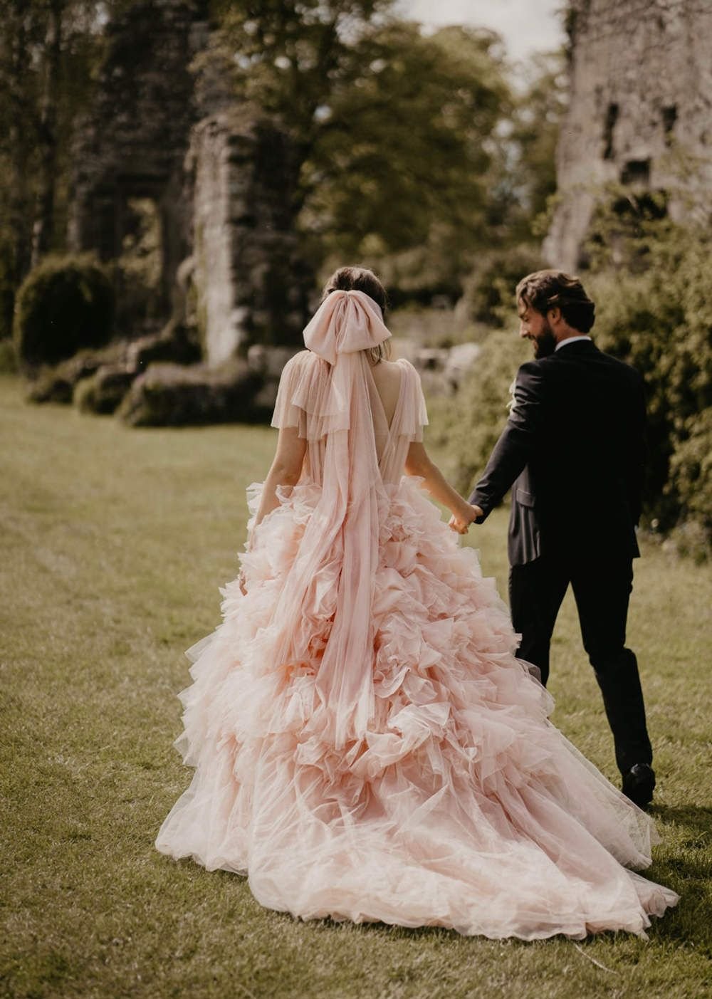 A couple walks hand-in-hand through a grassy field, with stone ruins and greenery in the background. The woman wears a voluminous pastel pink tulle gown with ruffled layers and a large pink bow in her hair, while the man is dressed in a black suit. The scene exudes romance and whimsy, set in a historic outdoor location.