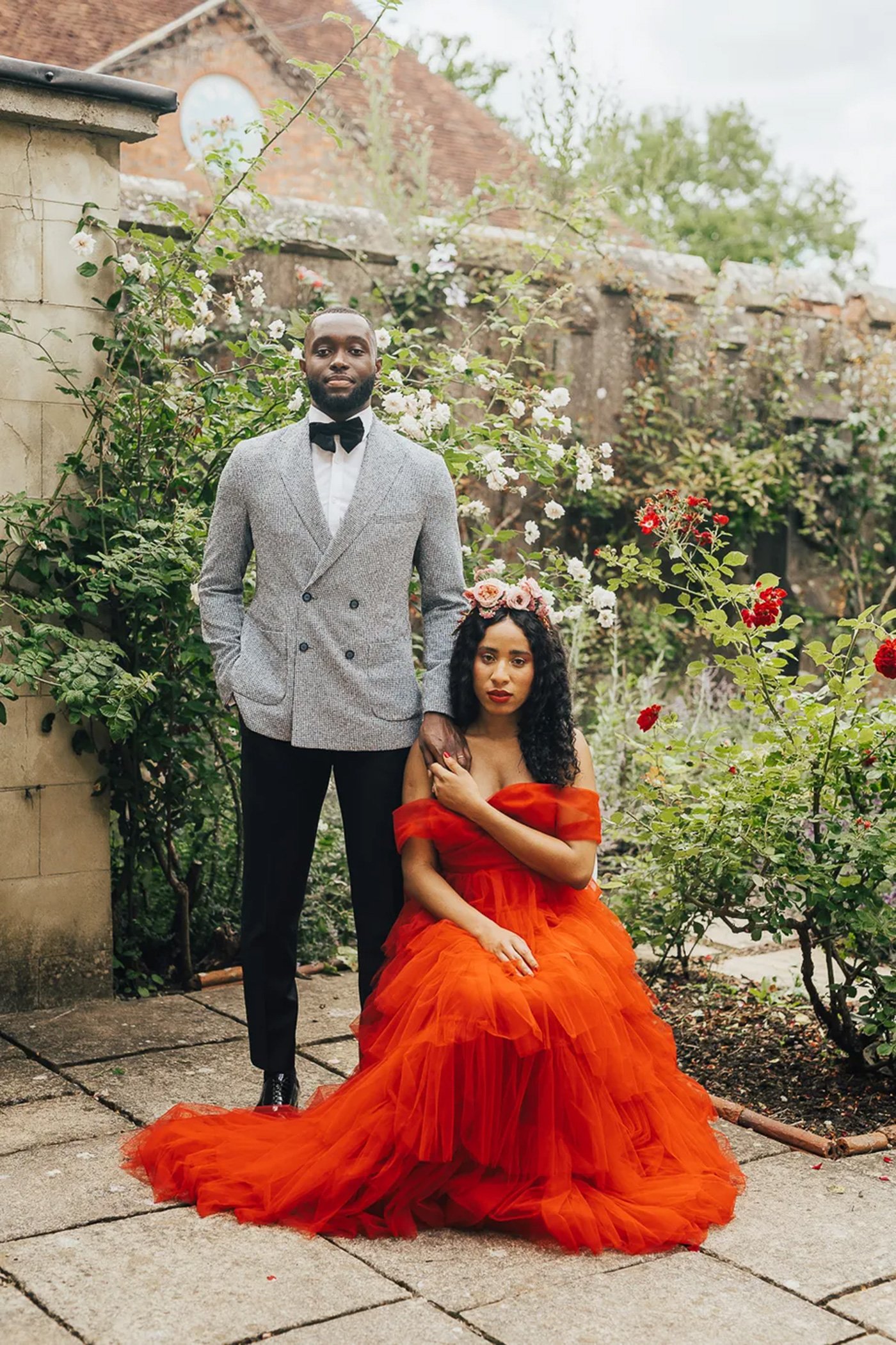 Um casal posa em um jardim cercado por rosas e folhagens. O homem está de pé, vestindo um terno cinza claro de duasfileiras com calças pretas e um laço preto. A mulher, sentada, usa um vestido vermelho fluido e uma coroa de flores. O ambiente é ao ar livre, perto de um muro de pedra coberto por flores.