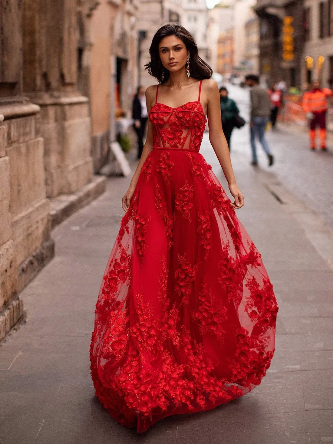 Una mujer camina con confianza por una calle empedrada con un vestido rojo vibrante con intrincados apliques florales. El vestido transparente tiene un corpiño ajustado con finos tirantes finos y una falda fluida y voluminosa. Su cabello oscuro y ondulado y unos llamativos pendientes completan su sofisticado look. El fondo difuminado muestra un paisaje urbano de estilo europeo con peatones y edificios históricos.