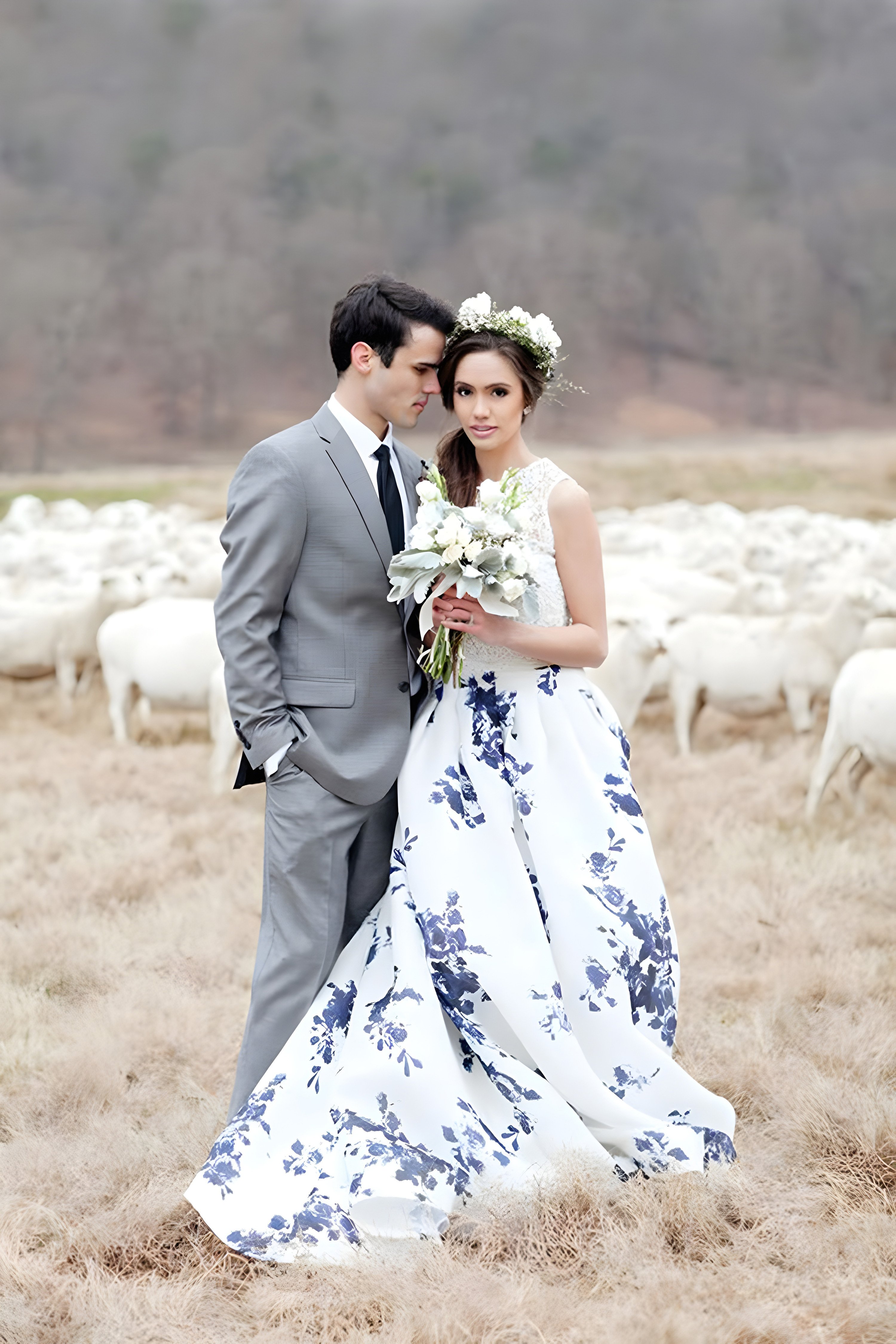 A bride in a white wedding gown with blue floral patterns stands in a field alongside the groom, who is dressed in a light gray suit. The bride holds a bouquet of white flowers and wears a flower crown. They are surrounded by a flock of sheep, and the landscape behind them is calm and rural.