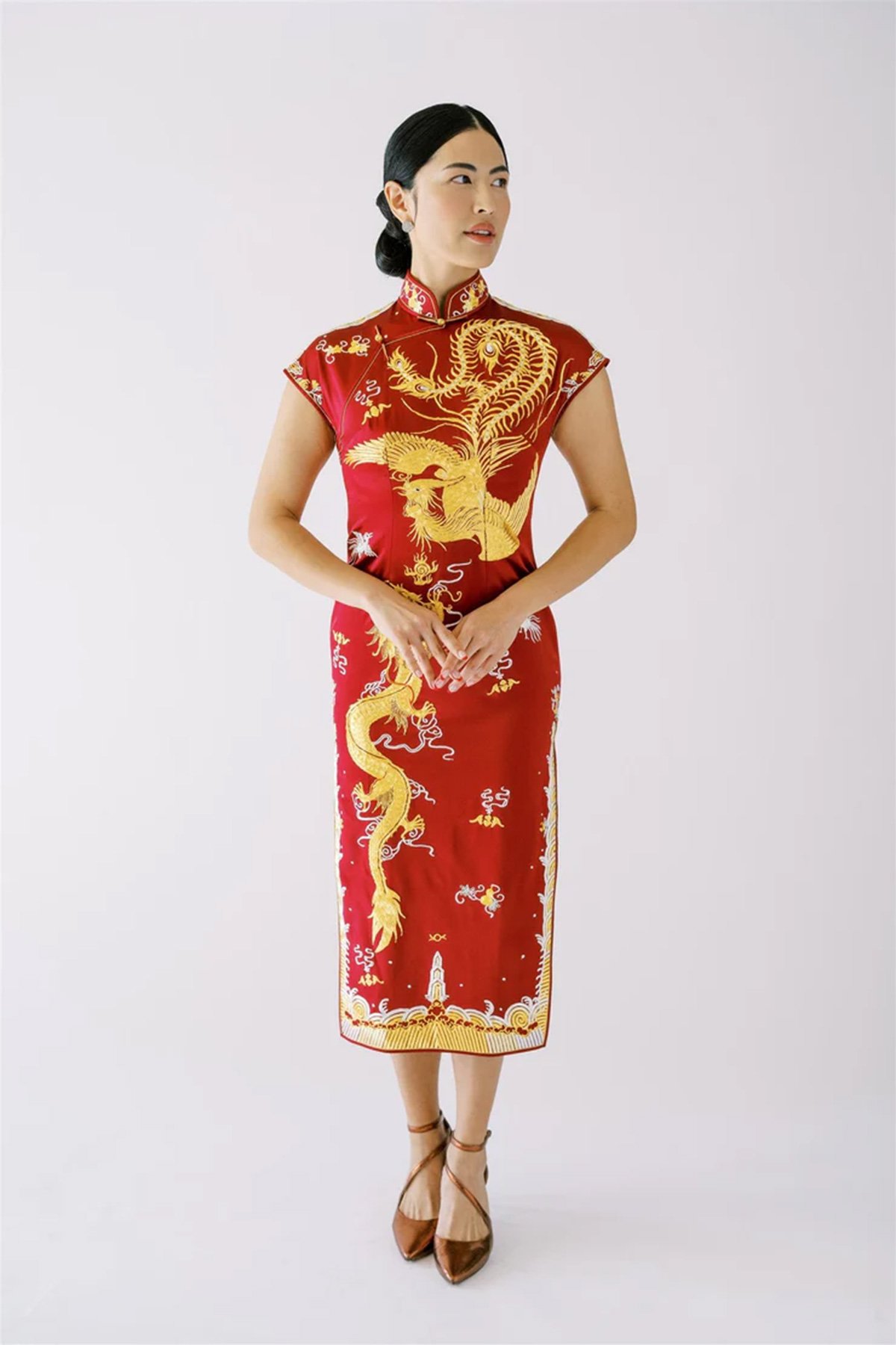 En kvinne står i en rød tradisjonell kinesisk brudekjole (cheongsam) med intrikate gullbroderier med dragemønster. Hun har hendene foldet foran seg og ser til siden, med håret satt opp i en lav knute. Bakgrunnen er helt hvit.