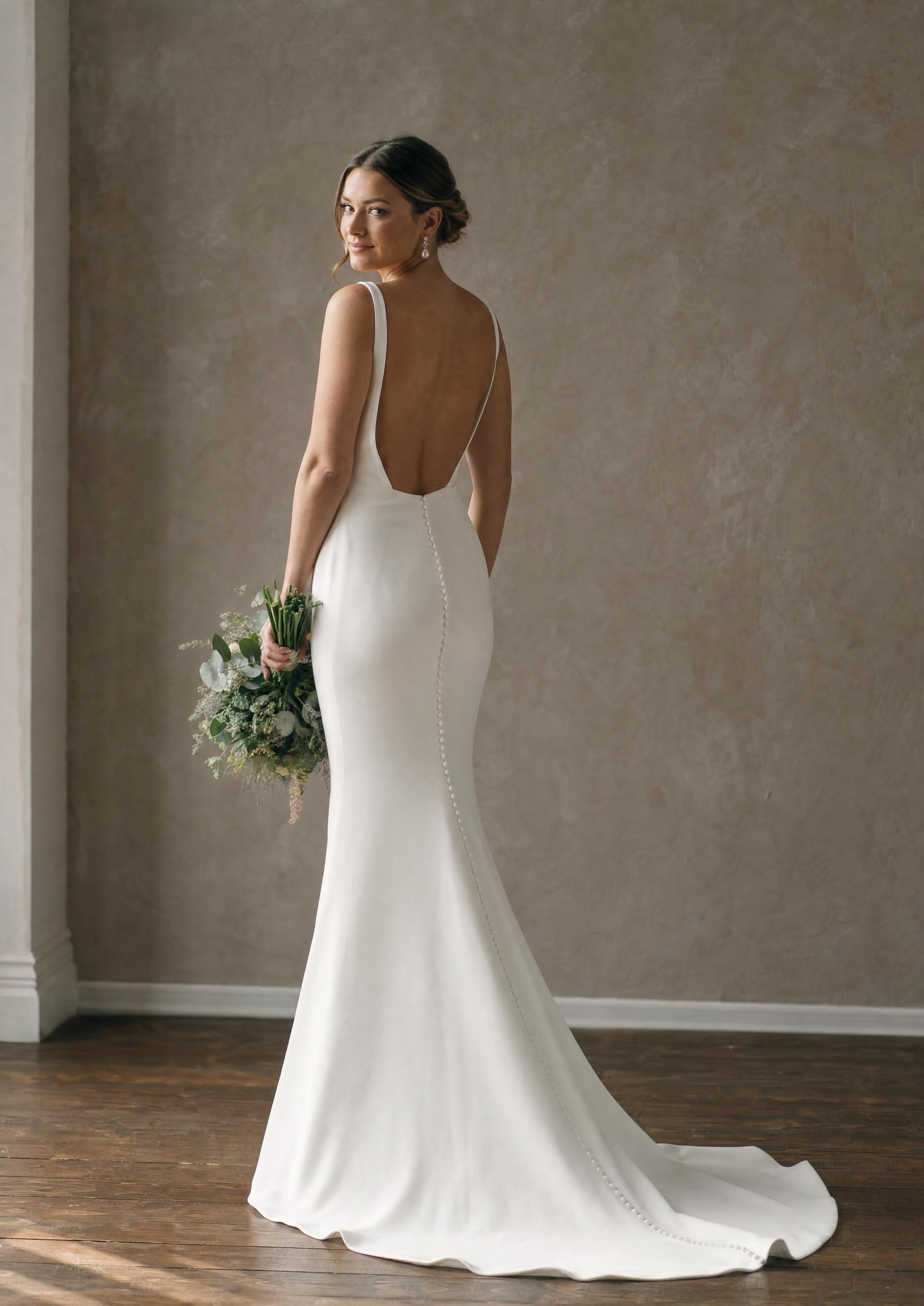 Mireasă într-o rochie de mireasă elegantă, din ivoire, care urmărește linia corpului, cu spatele ușor decupat, privind încrezătoare în lumina naturală difuză
