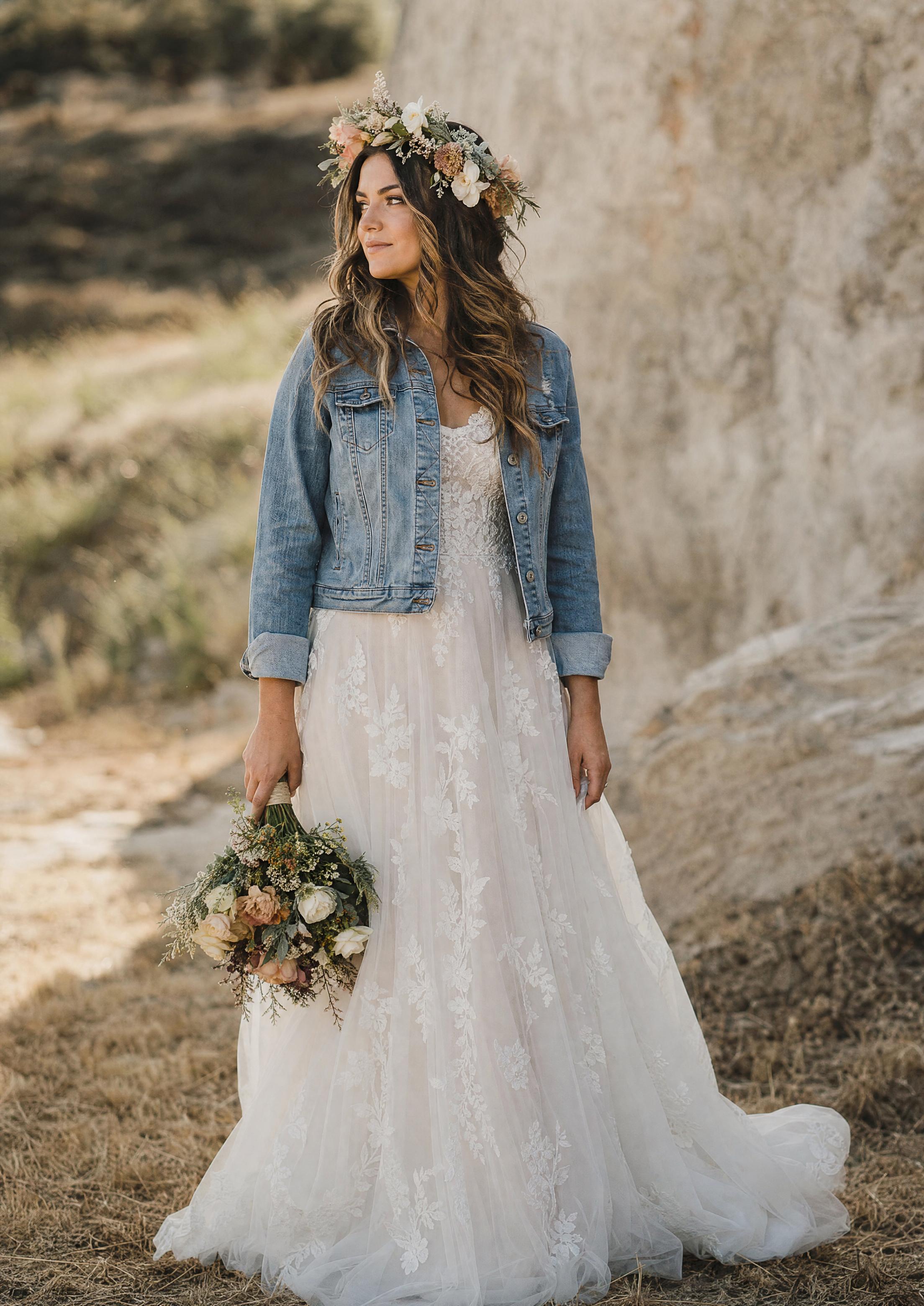 Núvia boho estilitzada amb un toc floral fresc al cabell, ones soltes i una jaqueta texana relaxada sobre un vestit fluid de puntes