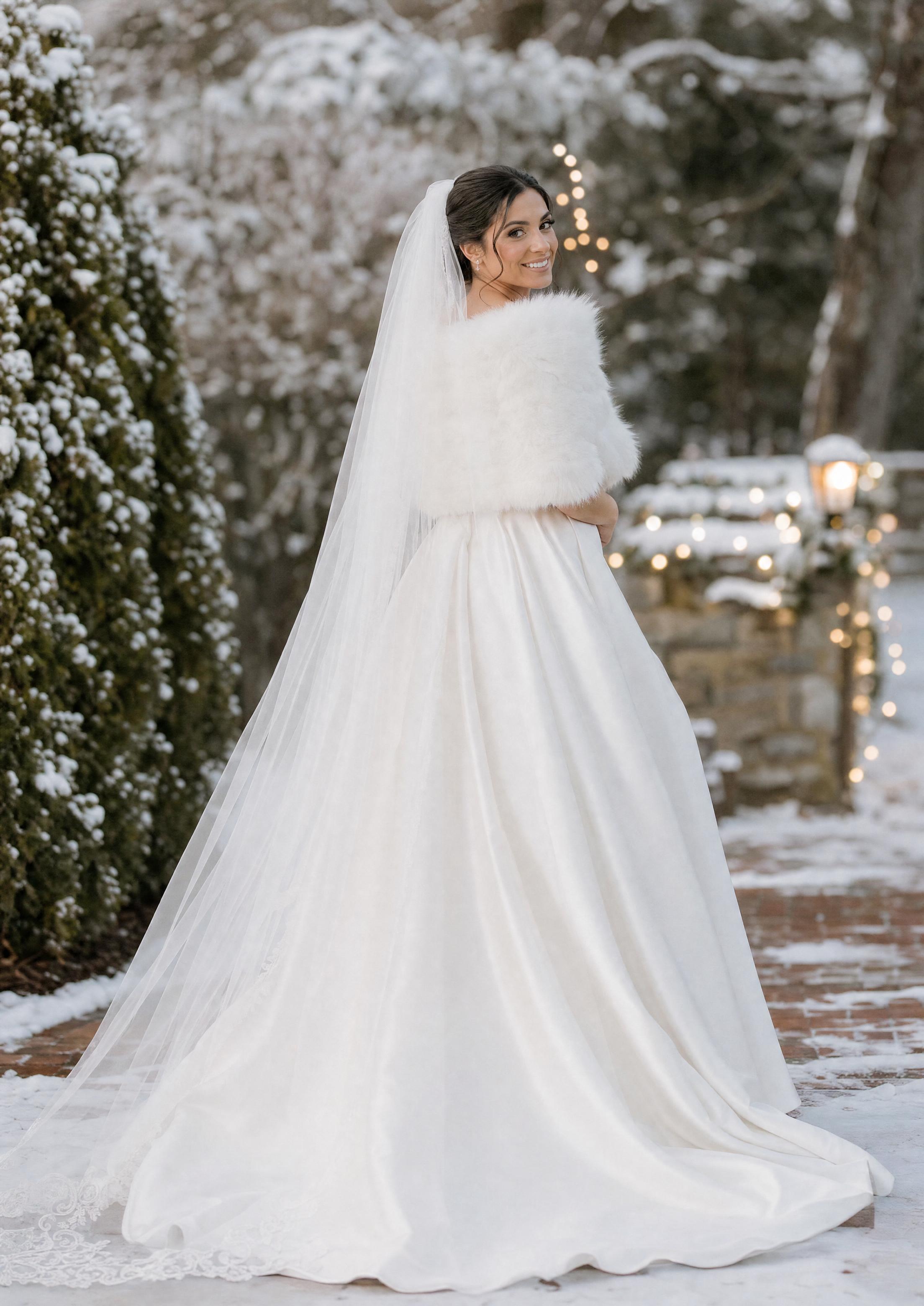 Núvia amb un vestit de núvia d’hivern amb una estola de pell sintètica i un vel llarg, posant en un entorn nevat i festiu
