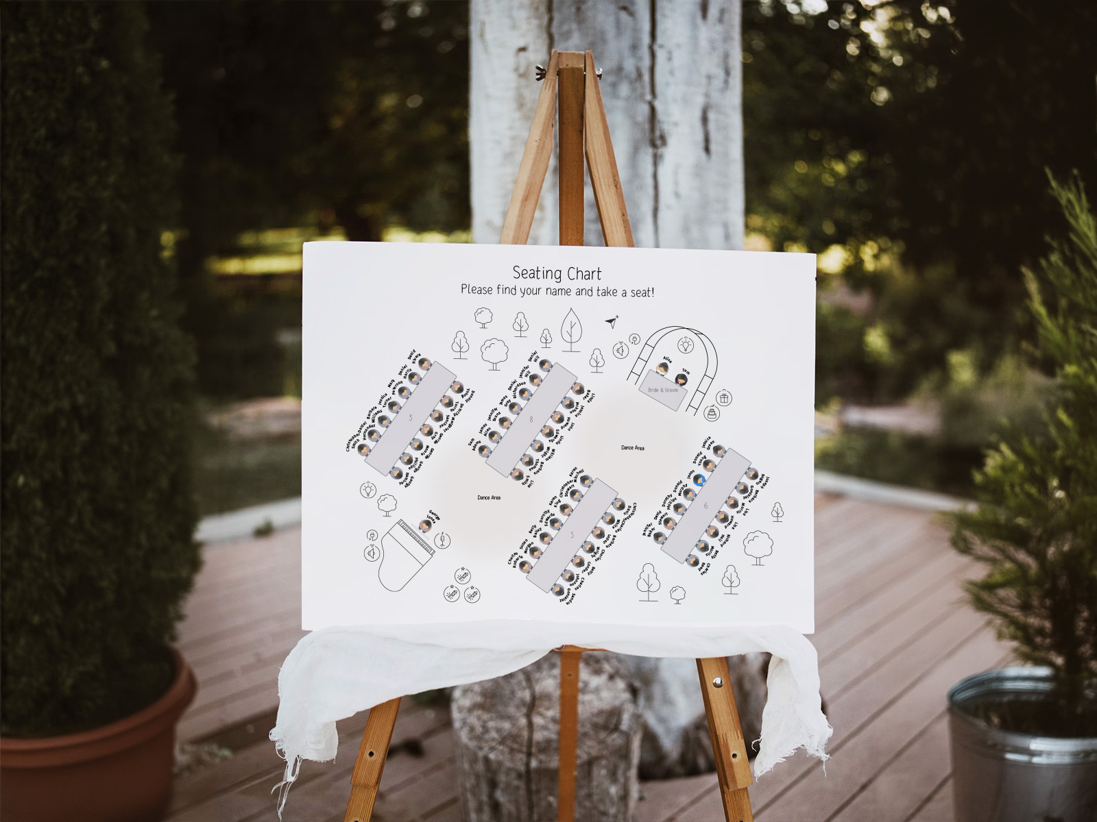 ගස් සහිත එළිමහන් උද්යාන ස්ථානයක ලී easel එකක විවාහ ආසන සැලැස්ම පෝස්ටරයක්, මධ්යම නර්තන ප්රදේශයක් වටා සකස් කළ දිග භෝජන මේස පහක් පෙන්වමින්